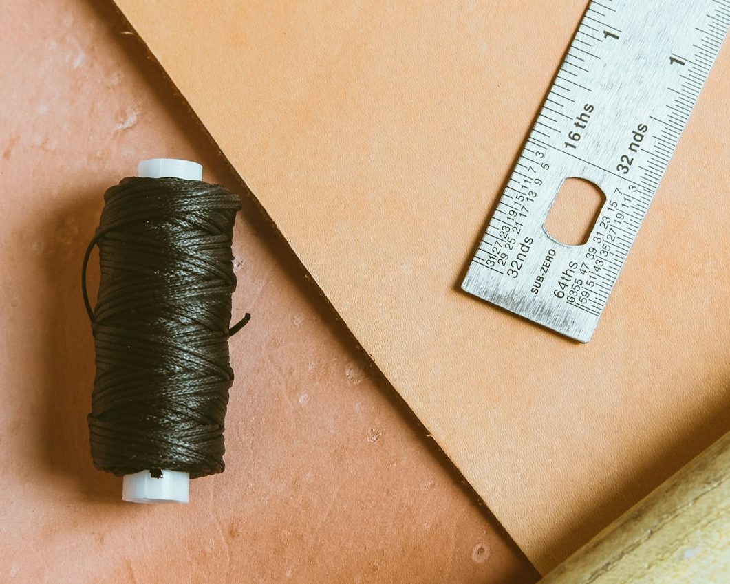 Dunkelbraunes Garn, orangefarbener Papierbogen und ein Metalllineal auf Leder.